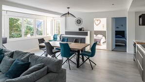 Een moderne woonkamer met een bank, eettafel en stoelen. Er is een keuken met een aanrecht en kasten. De kamer heeft grote ramen en uitzicht op groen.