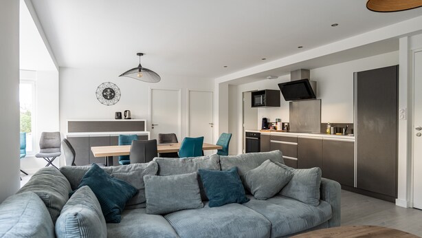 Een moderne woonkamer met een hoekbank, een houten salontafel en een keukenhoek met een flatscreen televisie en roestvrijstalen apparatuur.