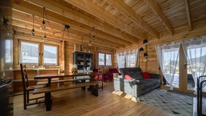 Interior de una cabaña de madera con comedor, sofá y amplias ventanas.