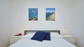 A neatly made bed with a white sheet and two blue pillows, a nightstand with a lamp, and two framed pictures of coastal scenes on the wall.