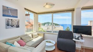 A modern living room with a sofa, armchair, and a small table with a TV. Large windows offer a view of the sea and buildings.