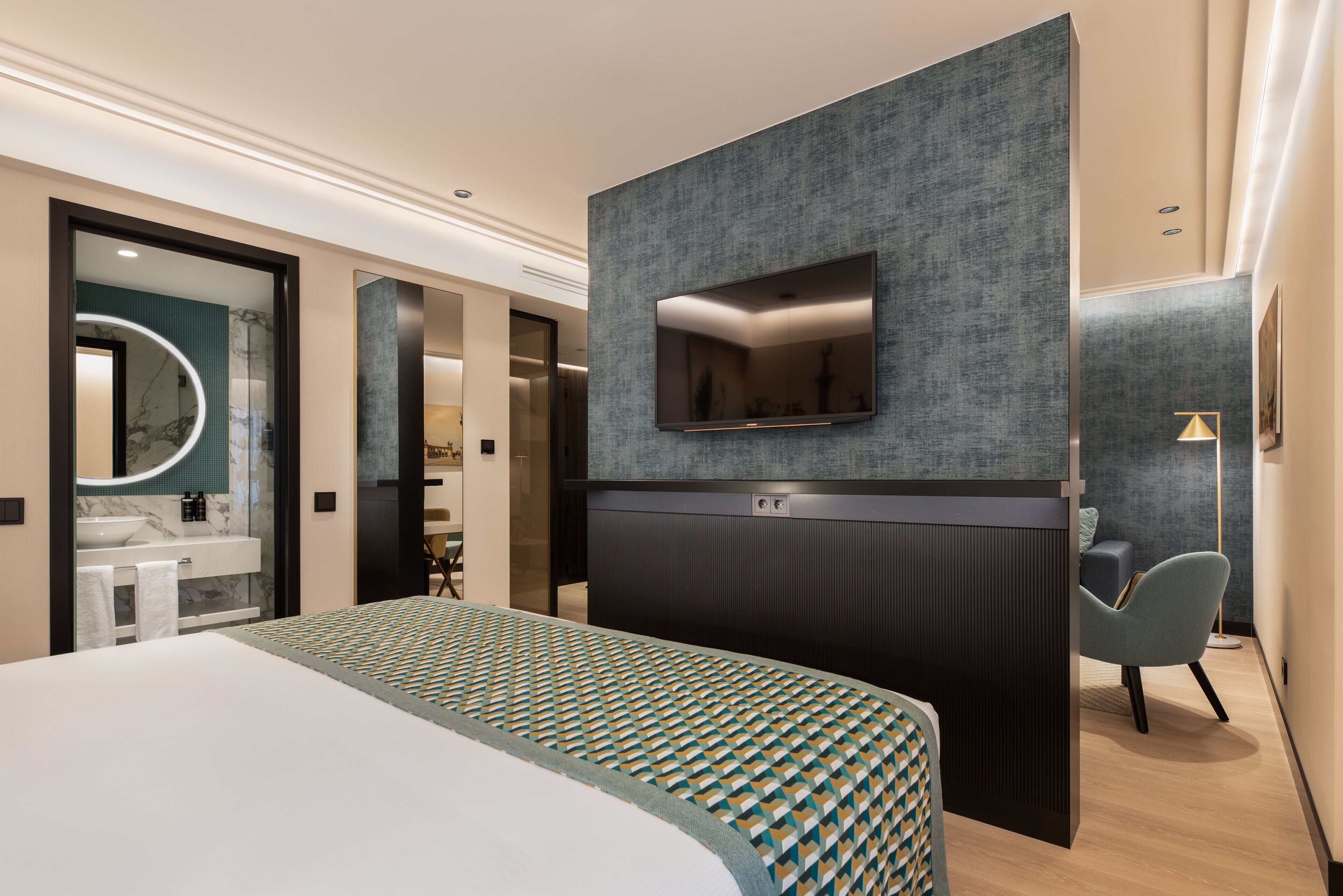 Una camera d'albergo moderna con un letto grande, una TV a parete, un bagno con lavabo in marmo e un'area salotto con una poltrona.