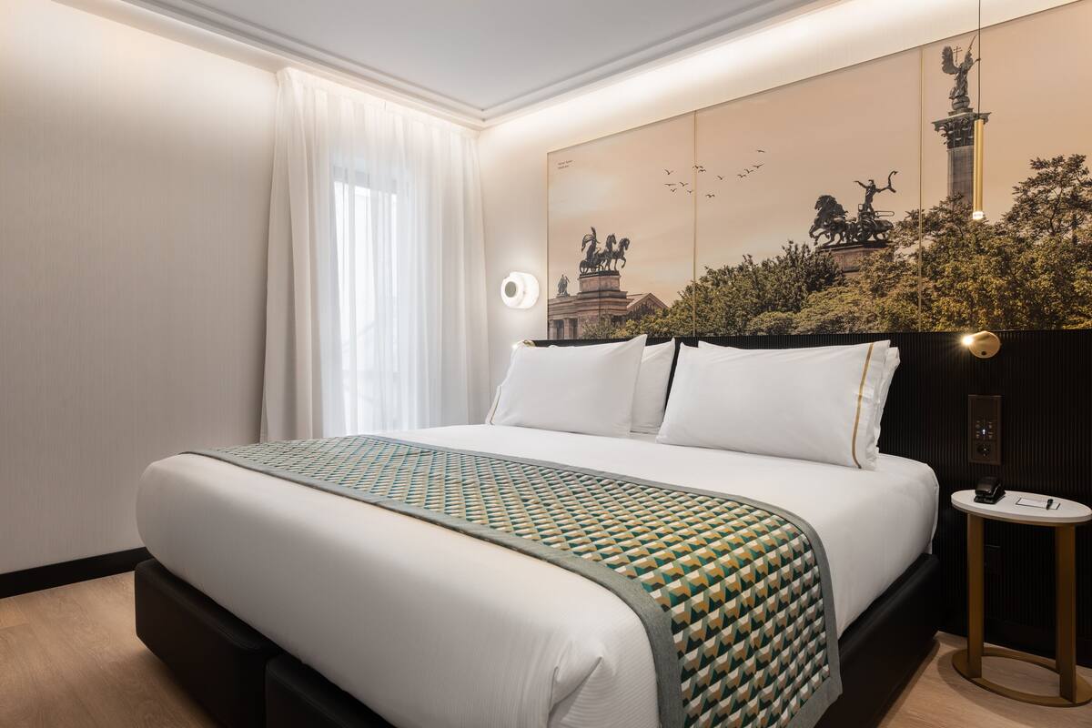 A hotel room with a large bed, a bedside table, a wall-mounted light, and a decorative wall piece.