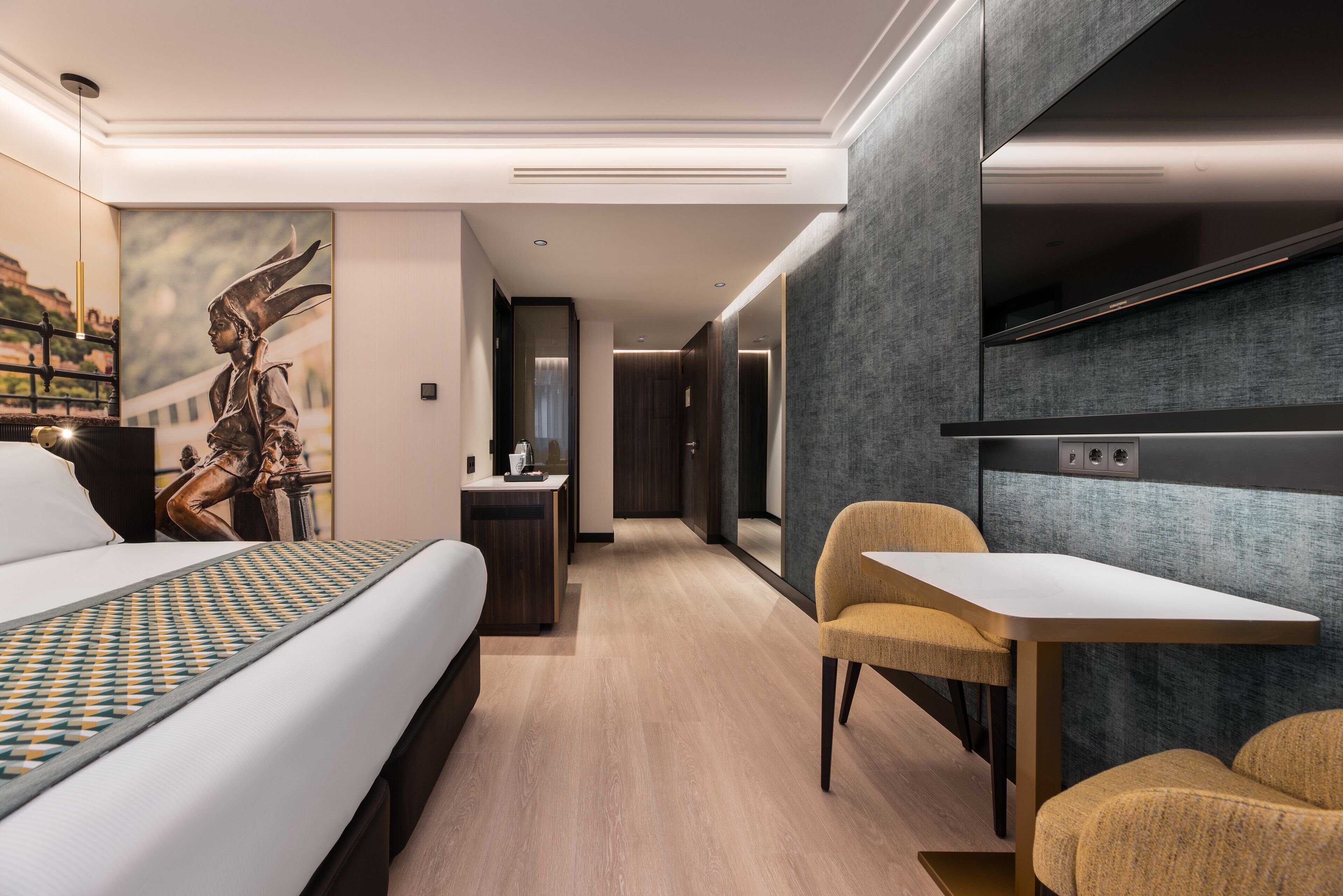 A modern hotel room with a bed, a desk, and a chair. There is a painting on the wall and a statue on the bed.