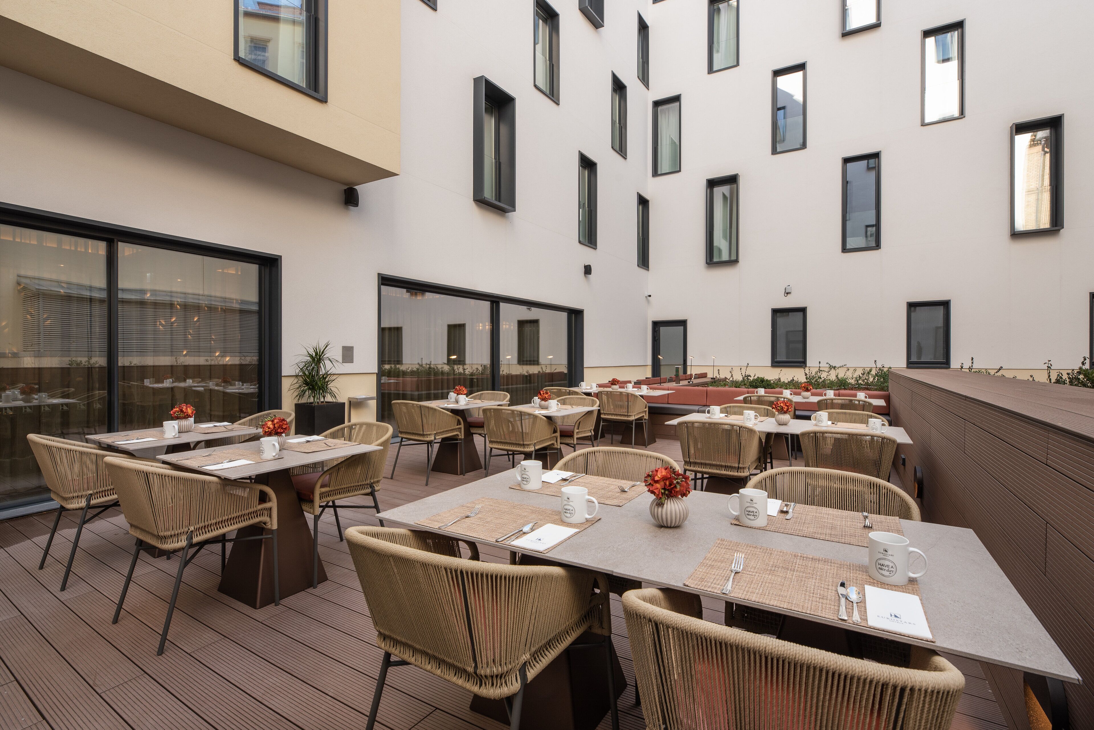 An outdoor dining area with multiple tables and chairs, large windows, and a modern building facade.