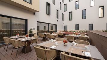An outdoor dining area with multiple tables and chairs, large windows, and a modern building facade.