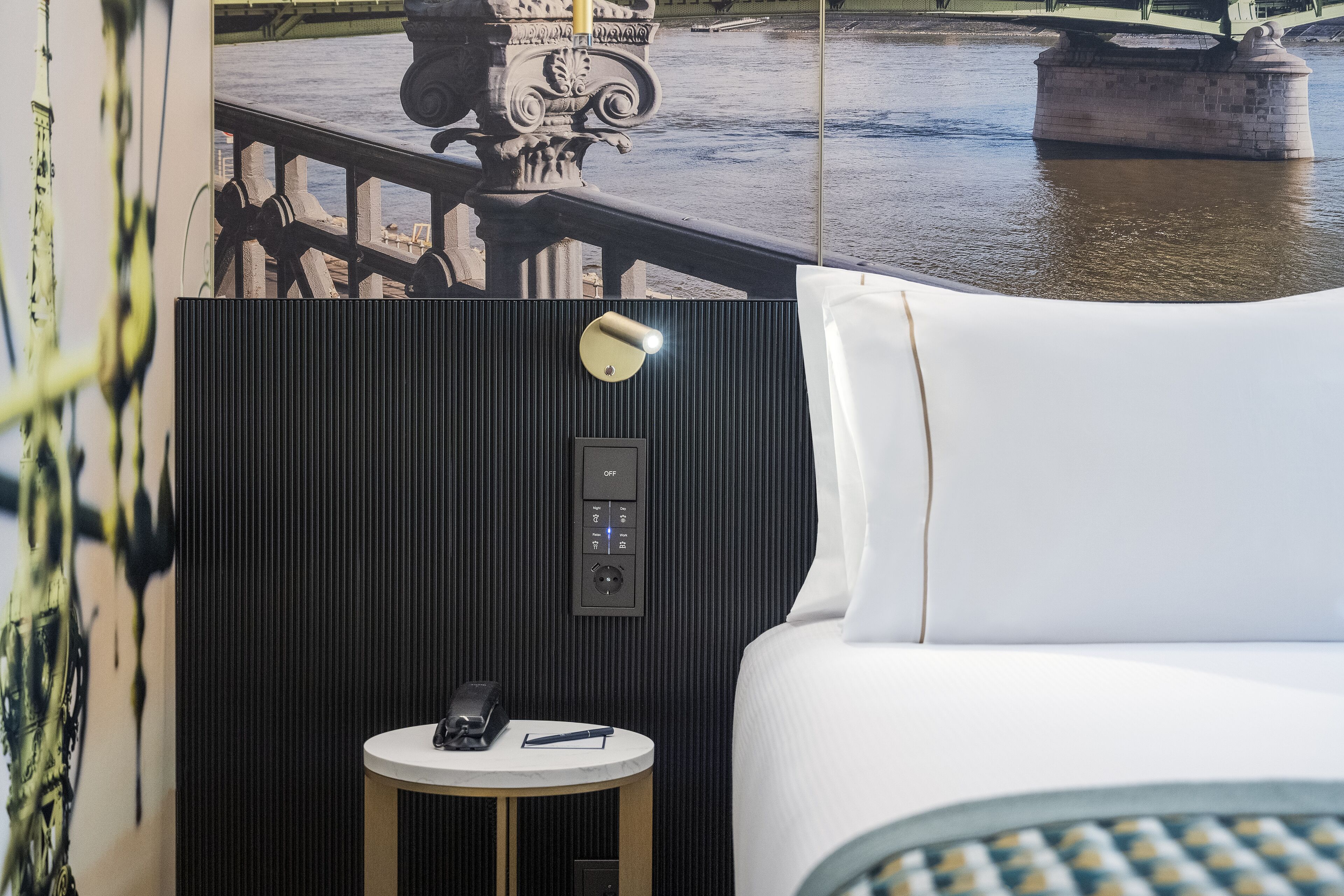 A hotel room with a view of a bridge and water, a control panel, a bedside table with a phone, and a bed with white linens.
