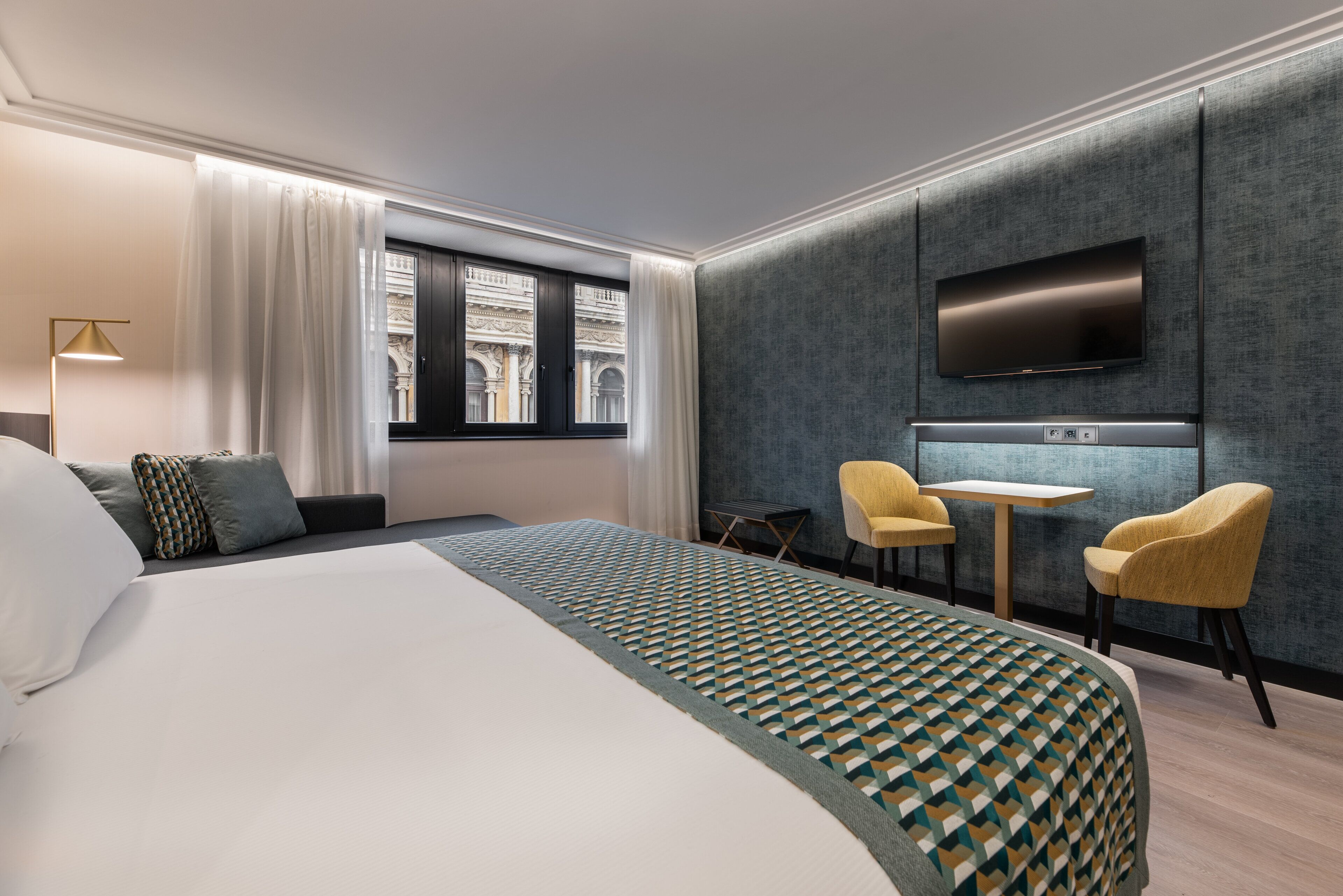 Habitación de hotel con una cama grande, un escritorio con dos sillas, un televisor de pantalla plana y un ventanal con cortinas.