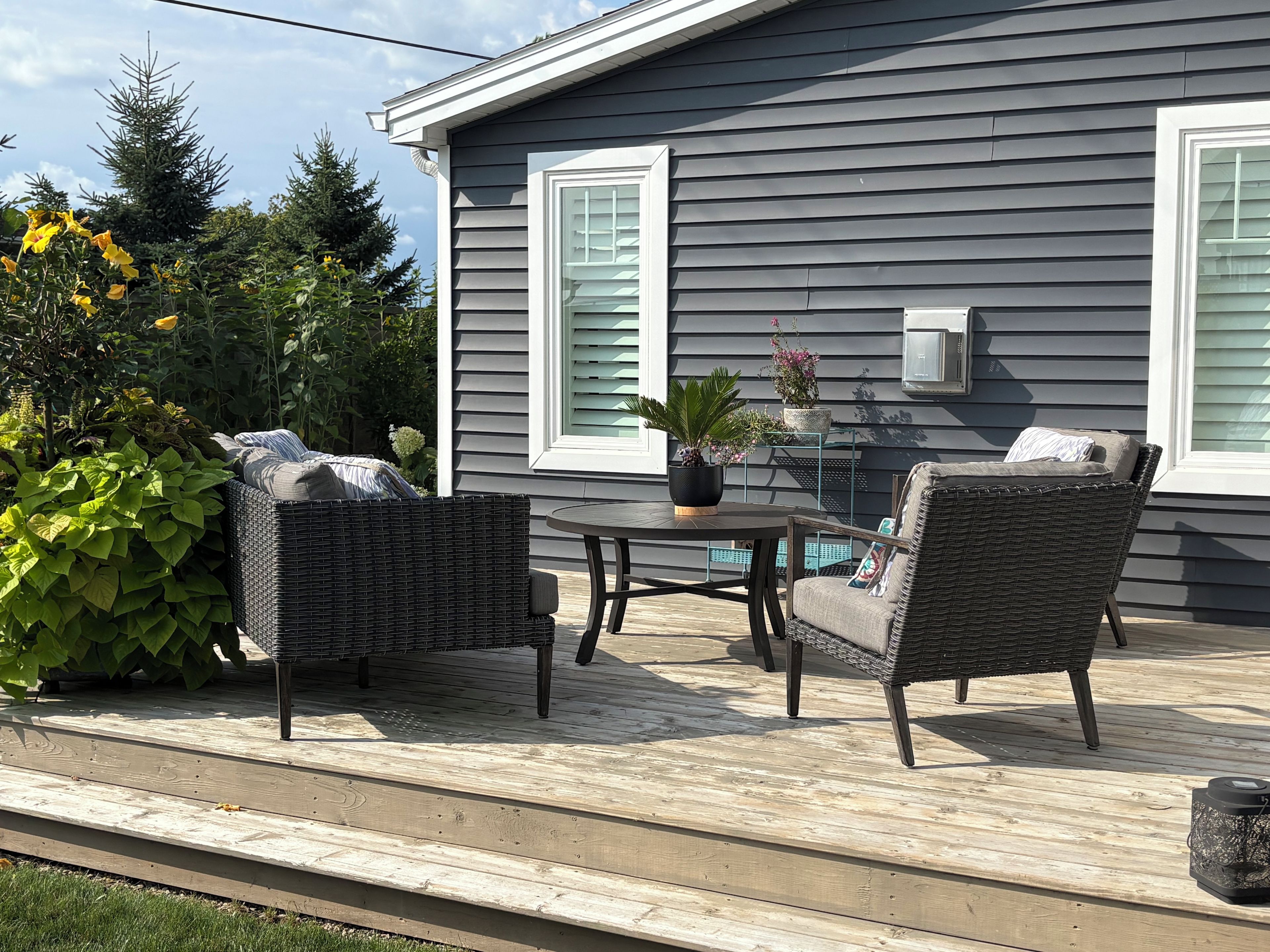 A deck with wicker furniture and a potted plant, adjacent to a house with gray siding and white trim.