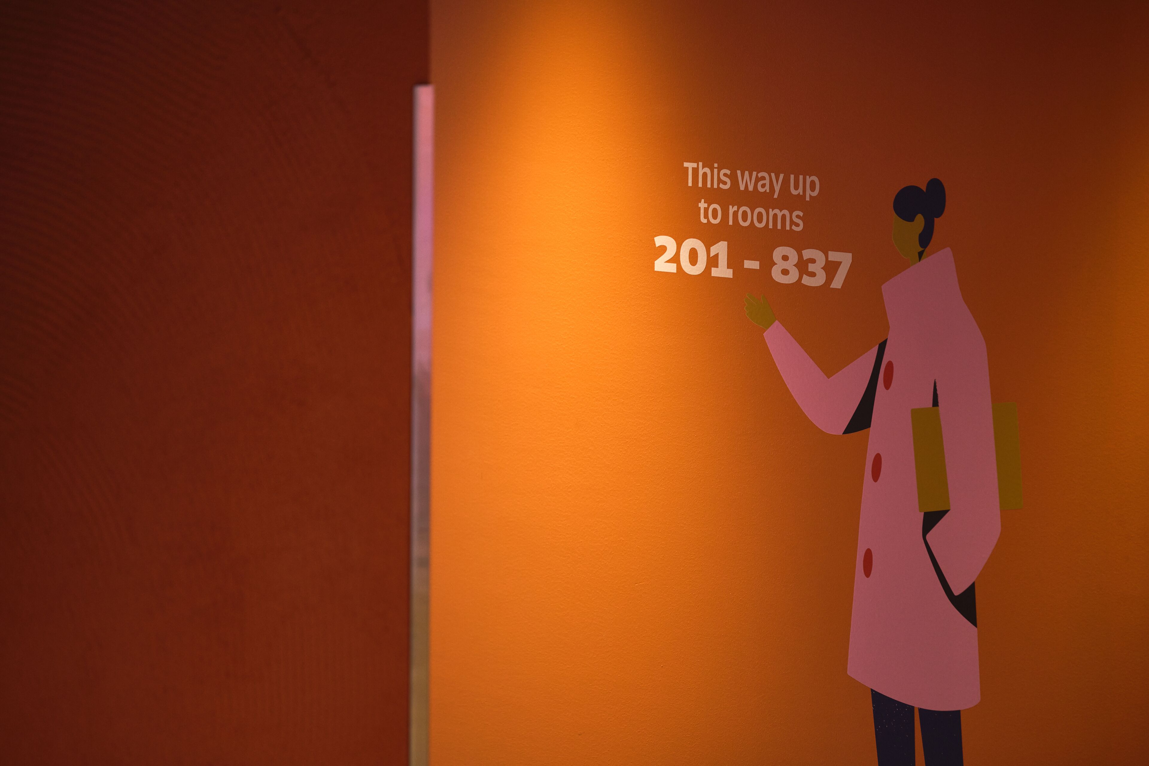A person in a pink coat pointing to a sign with room numbers 201-837.