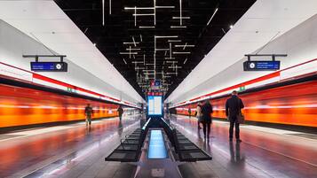En moderne togstation med et centralt perron, skilte over hovederne og et orange tog på sporet.