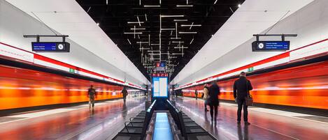 Una estación de tren moderna con andén central, indicaciones colgantes y un tren de color naranja brillante sobre las vías.
