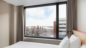 A hotel room with a large window, a bed with white linens, and a view of trees and buildings outside.