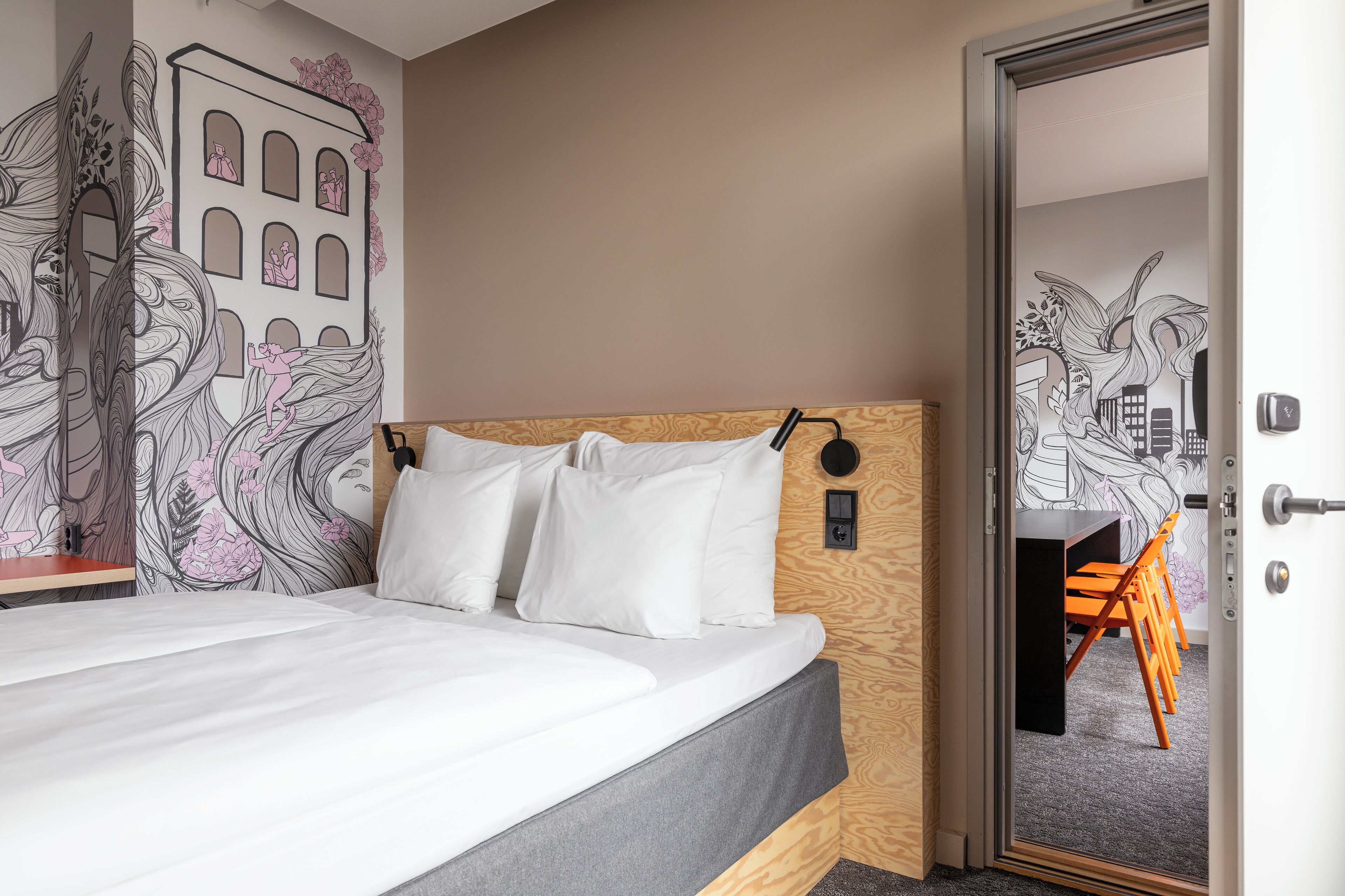 A bedroom with a bed, white pillows, a wooden headboard, and a door leading to a room with a desk and chairs.