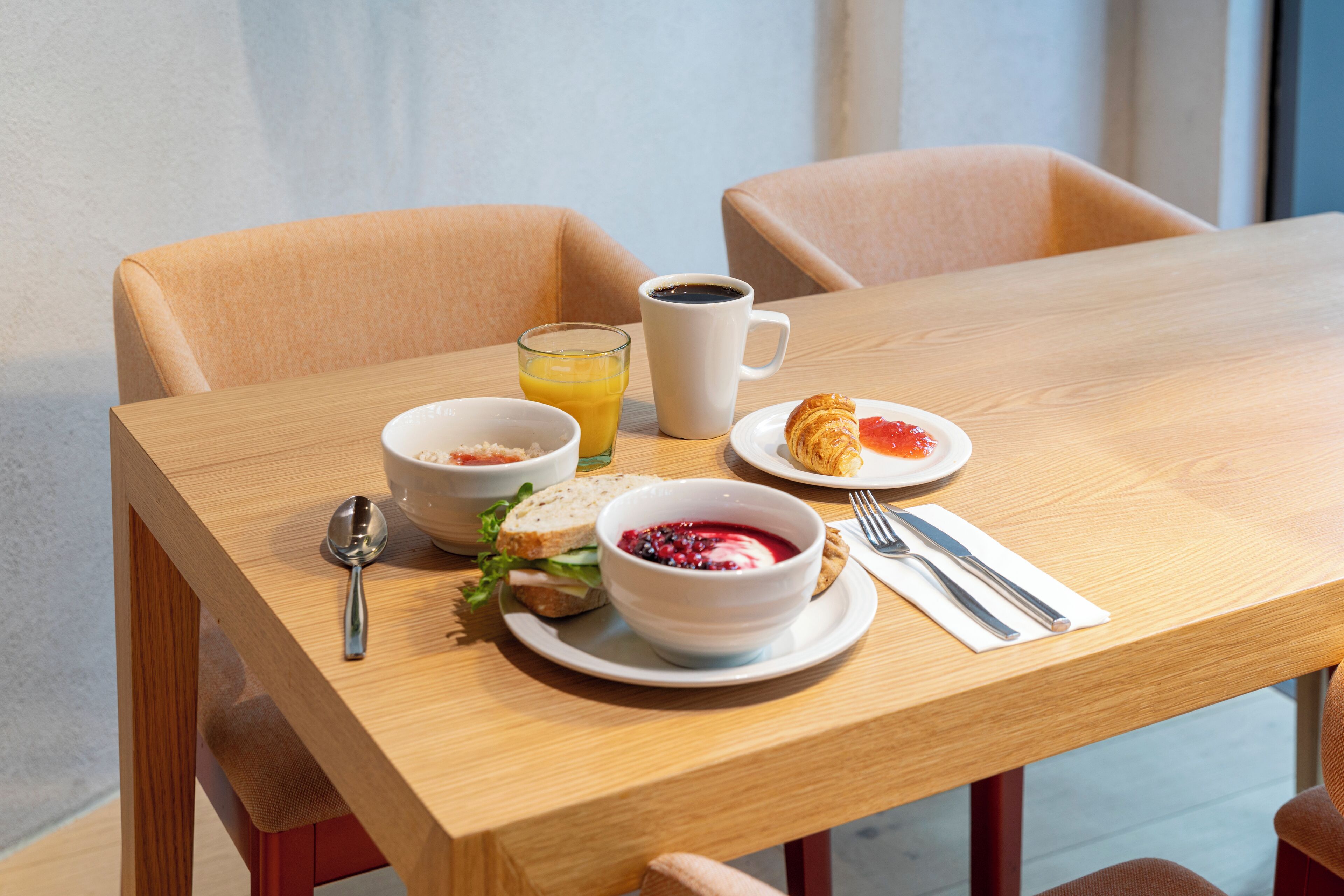 Una mesa de desayuno con un tazón de cereal, un vaso de jugo de naranja, una taza de café, un croissant y un sándwich.