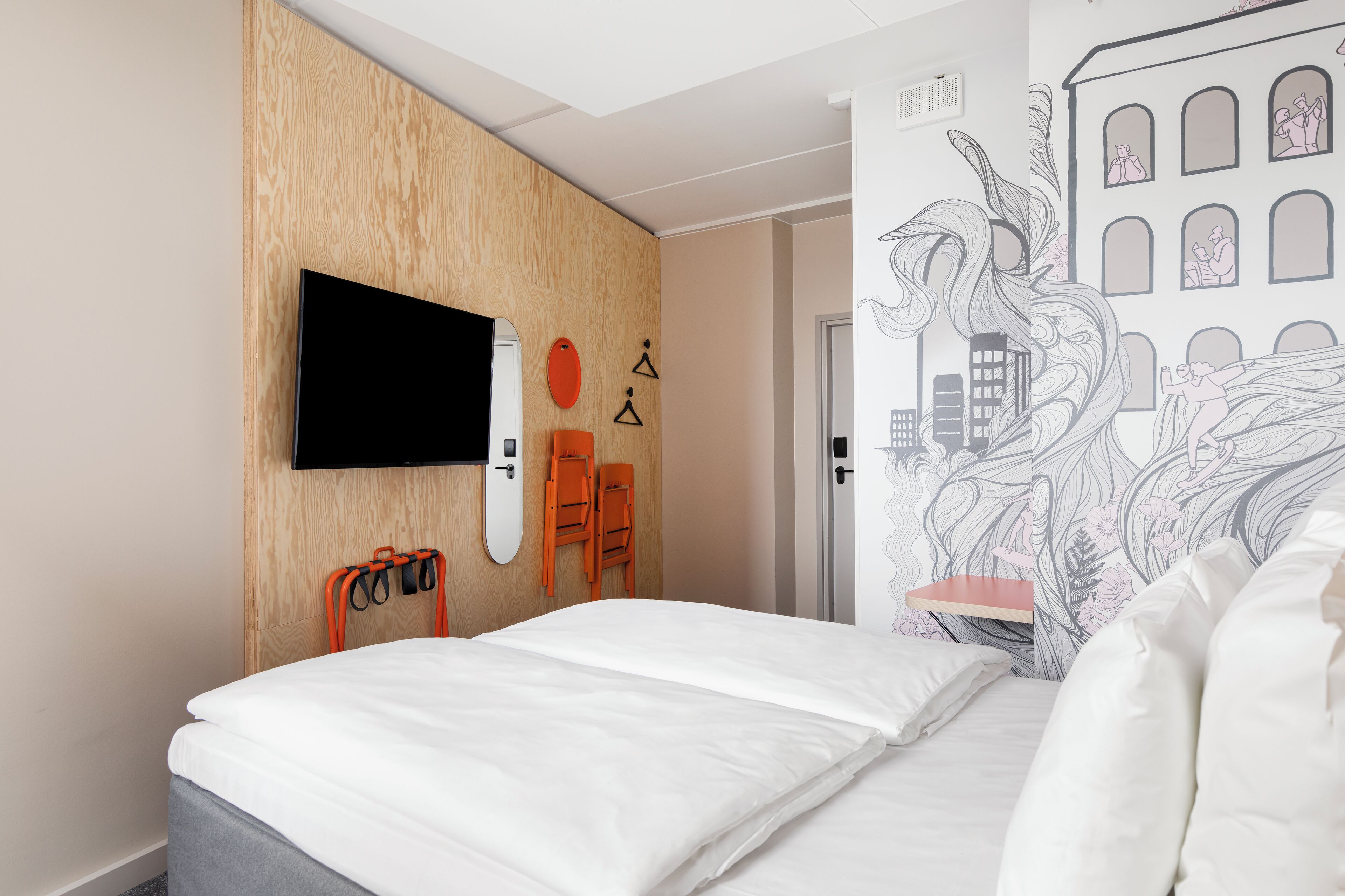 A hotel room with a bed, a TV mounted on the wall, a wooden panel design, and a door with a cityscape mural.