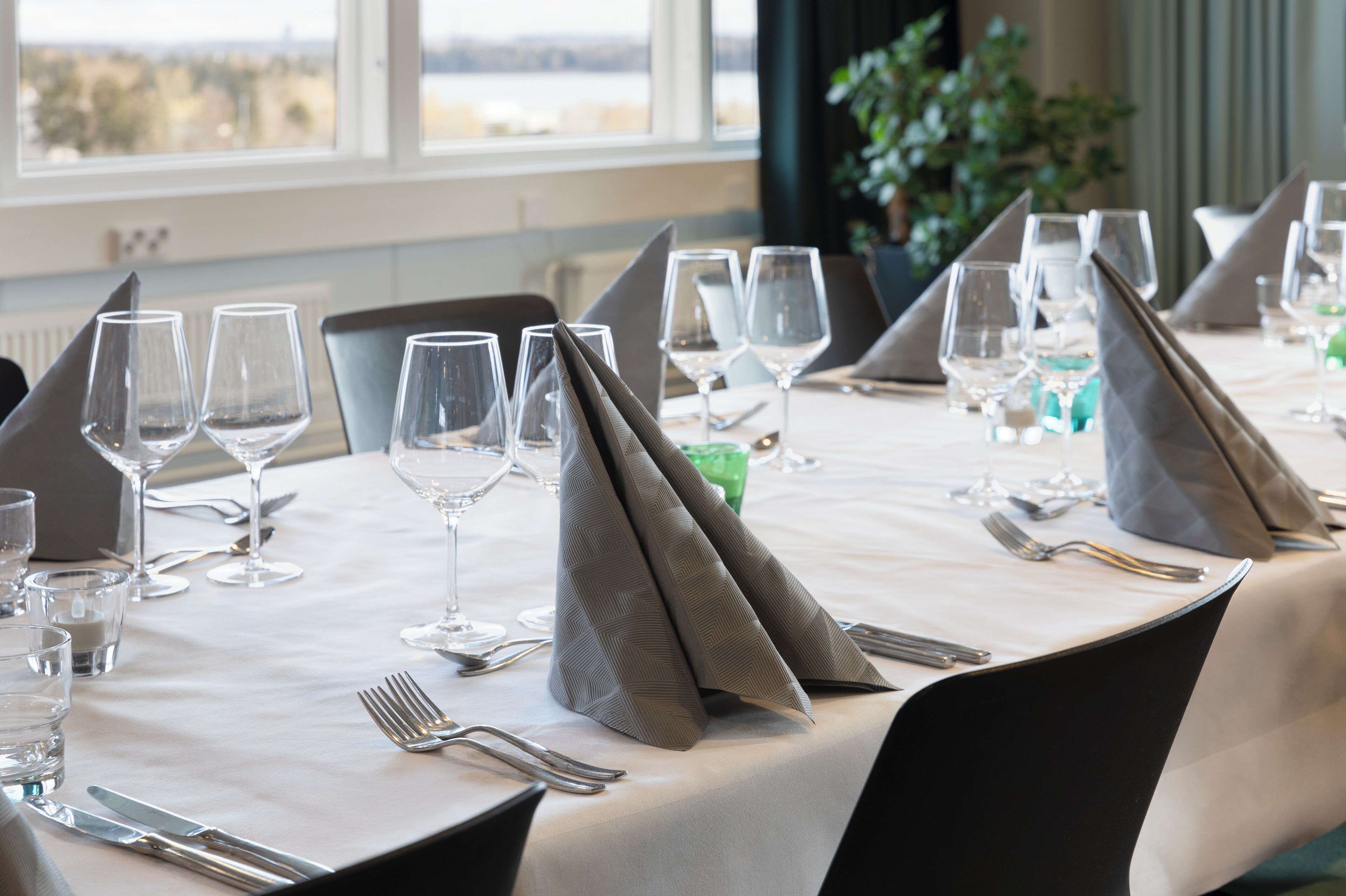 Ett matbord dukat med flera vinglas, bestick och vikta servetter, med utsikt över en stadssiluett genom stora fönster.