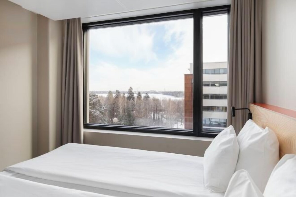 A hotel room with a large window, a bed with white linens, and a view of trees and buildings outside.