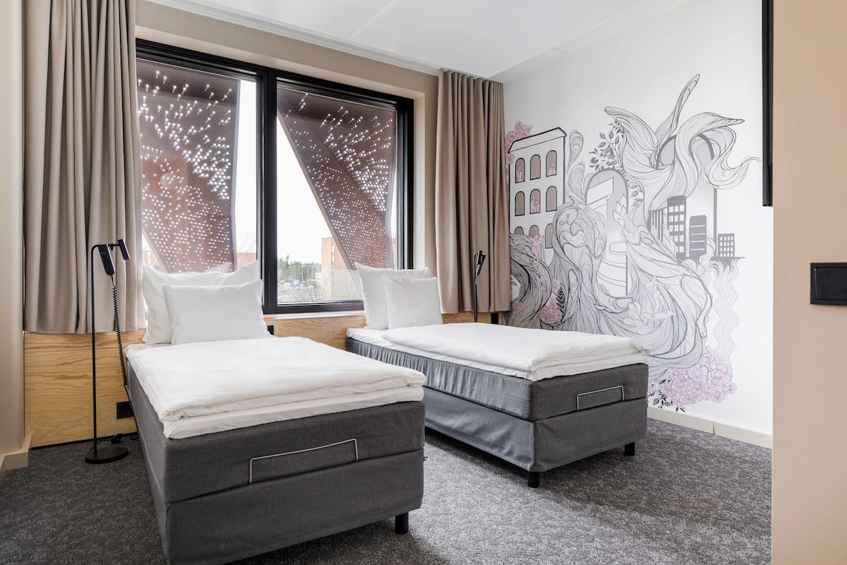 A hotel room with two beds, a large window with curtains, a cityscape mural, and a wooden floor.