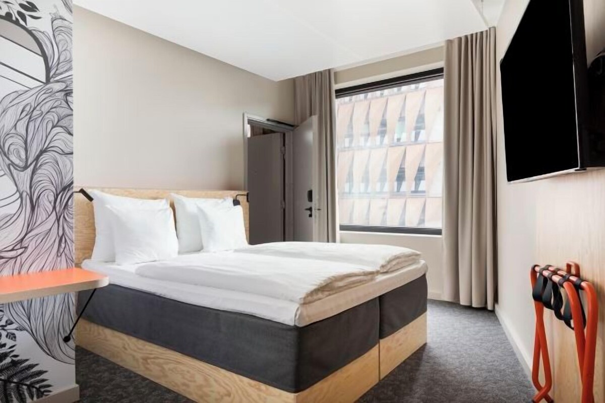 A hotel room with a bed, a television, a window with curtains, and a door.