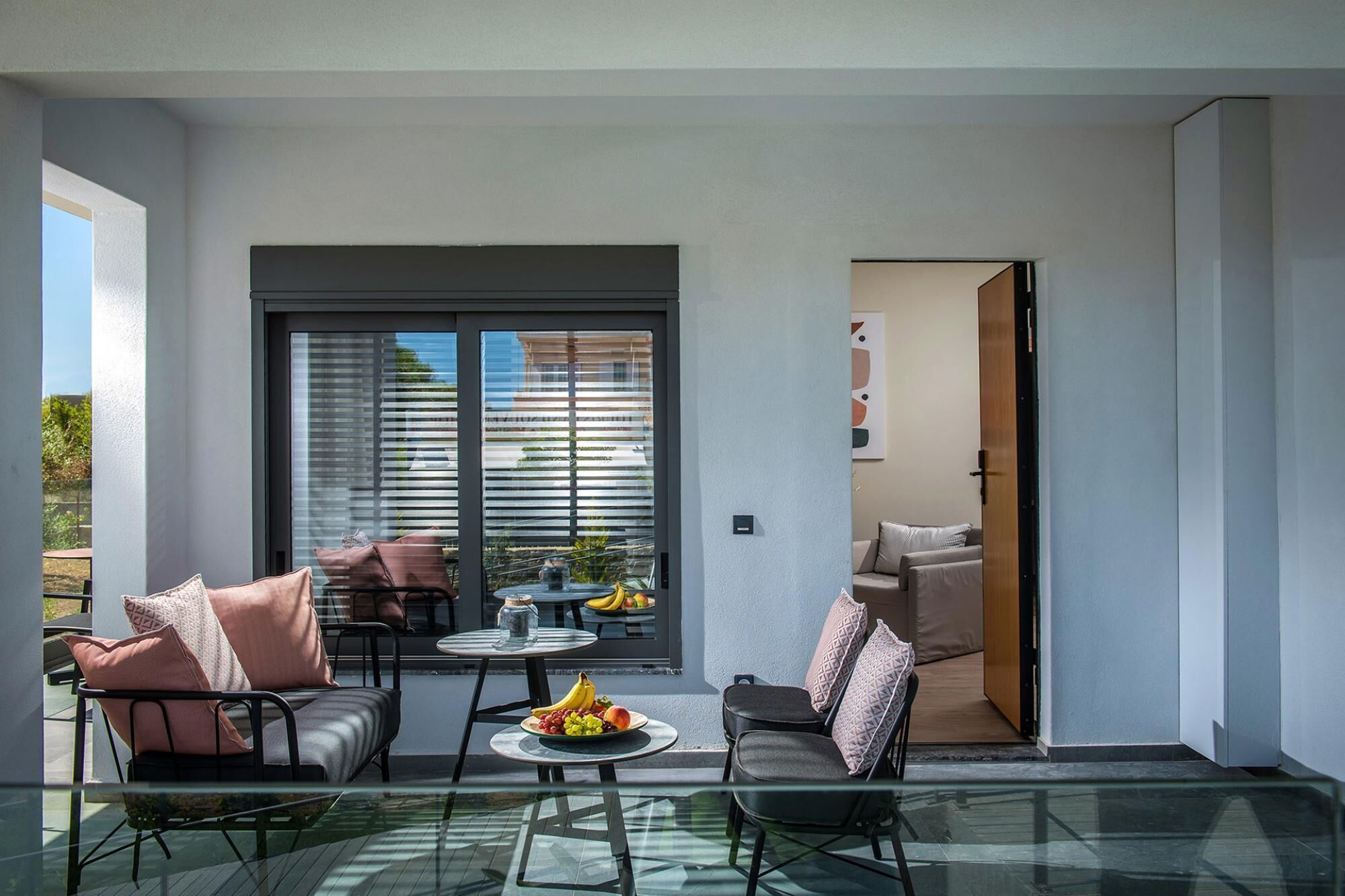 Un balcón moderno con una mesa de vidrio, dos sillas y una mesita redonda con fruta.