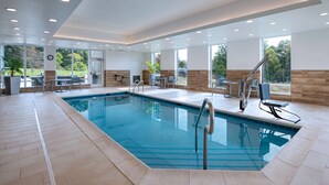 Una piscina interior moderna con paredes de vidrio transparente, suelo de baldosas y zona de estar con mesas y sillas.