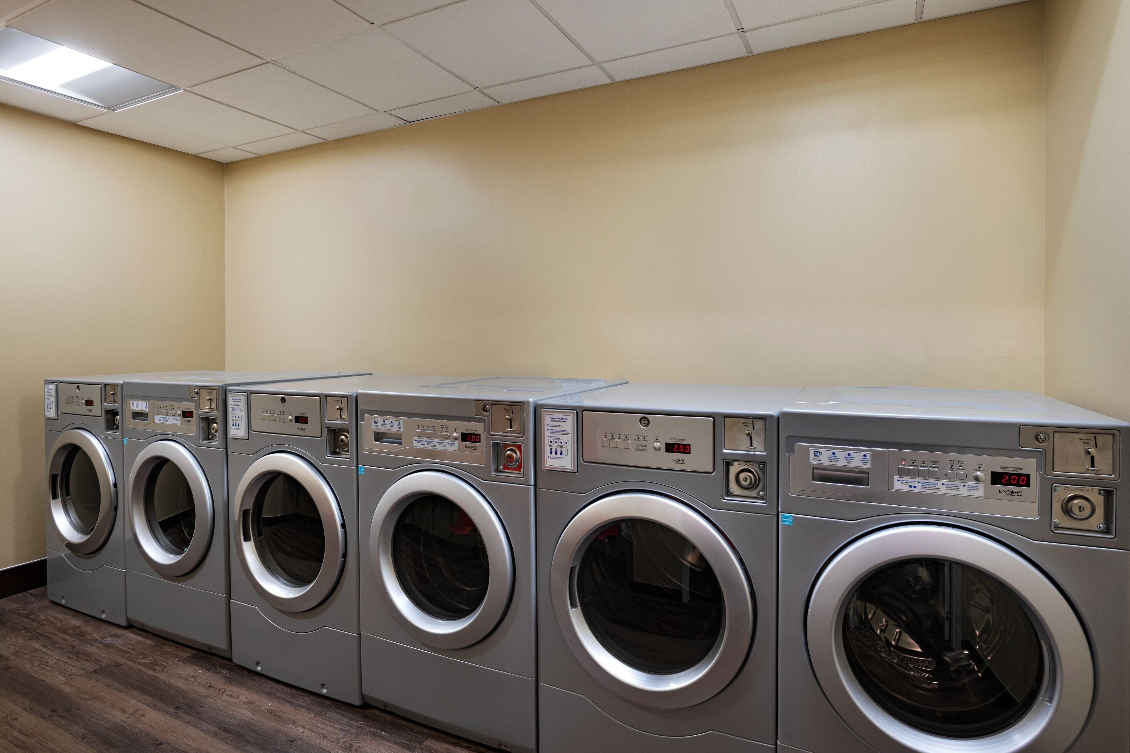 A row of front-loading washing machines in a laundry room with beige walls and a wooden floor.