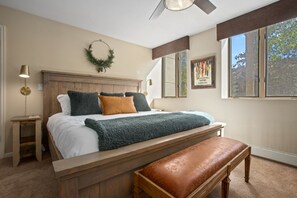 Une chambre avec un lit en bois, un banc, une couronne décorative accrochée au mur, un tableau et une fenêtre donnant sur des arbres.