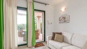 A living room with a white sofa, green curtains, a sliding glass door leading to an outdoor seating area, and a wall-mounted light fixture. - Domos Pintadas Apt. 4 Porto Rotondo (Olbia)