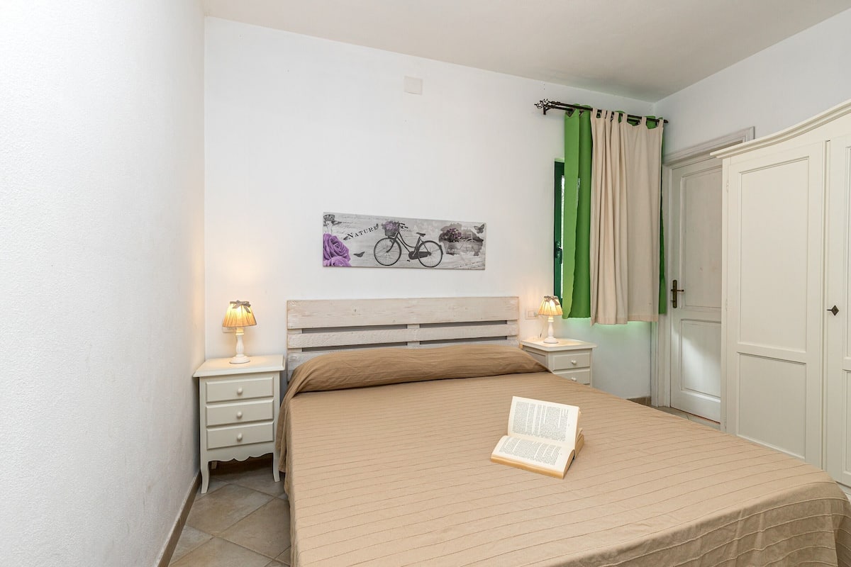 Quarto com cama, mesa de cabeceira, abajur, livro, obra de arte na parede, porta e cortina.