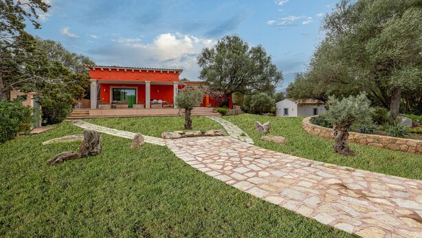 A red building with a stone pathway leading to it, surrounded by greenery and trees. - Domos Pintadas Apt. 4 Porto Rotondo (Olbia)