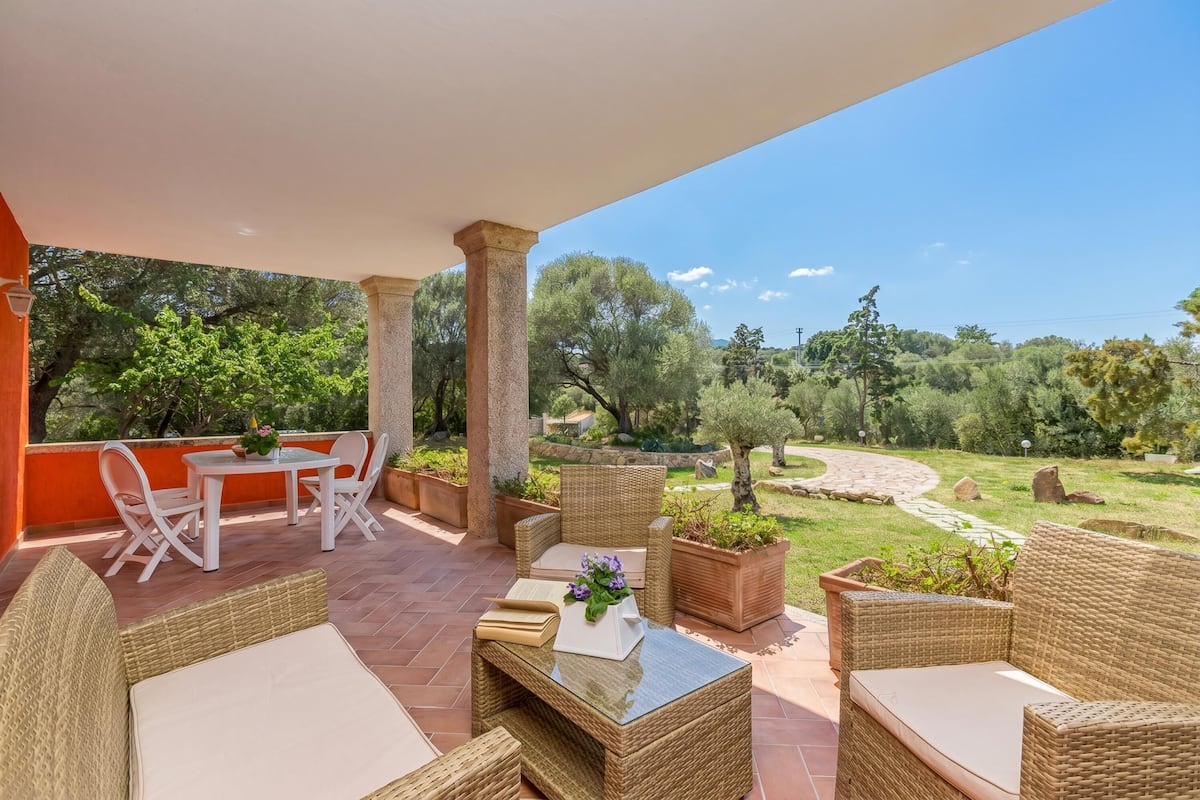 Alpendre coberto com móveis de vime, uma mesa com um vaso e vista para um jardim bem cuidado.