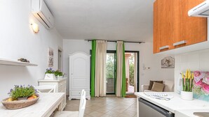 A modern kitchen with white cabinets, a dining table with a basket of fruit, and a view to the outside through a glass door. - Domos Pintadas Apt. 4 Porto Rotondo (Olbia)
