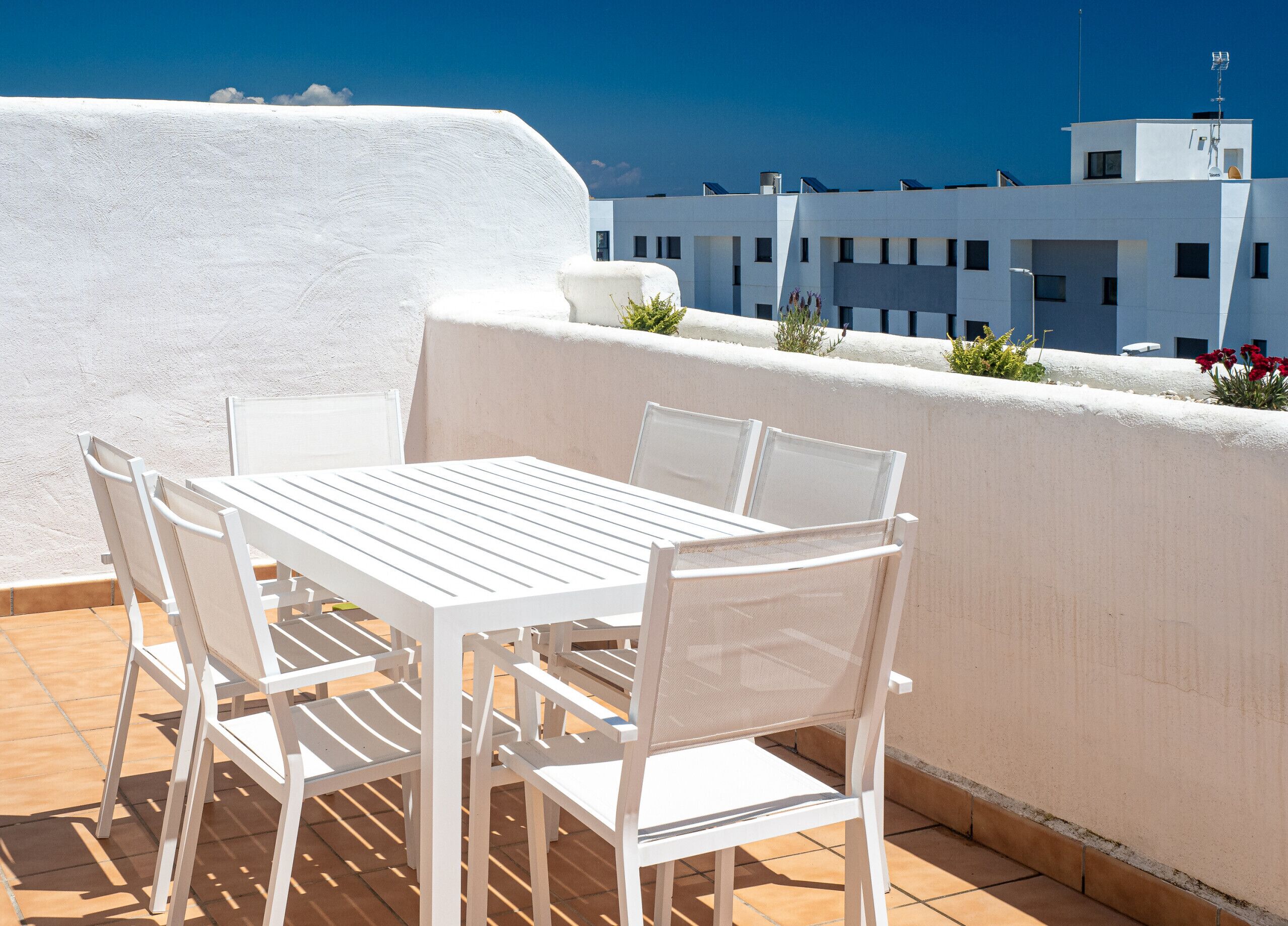 Una mesa y sillas blancas en una terraza con vistas a un edificio blanco y un cielo azul despejado.