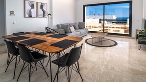 A modern living room with a wooden dining table, black chairs, a grey sofa, and a large window offering a view of the sea. - Lotus 2341 Golf beach & pool (Casares)