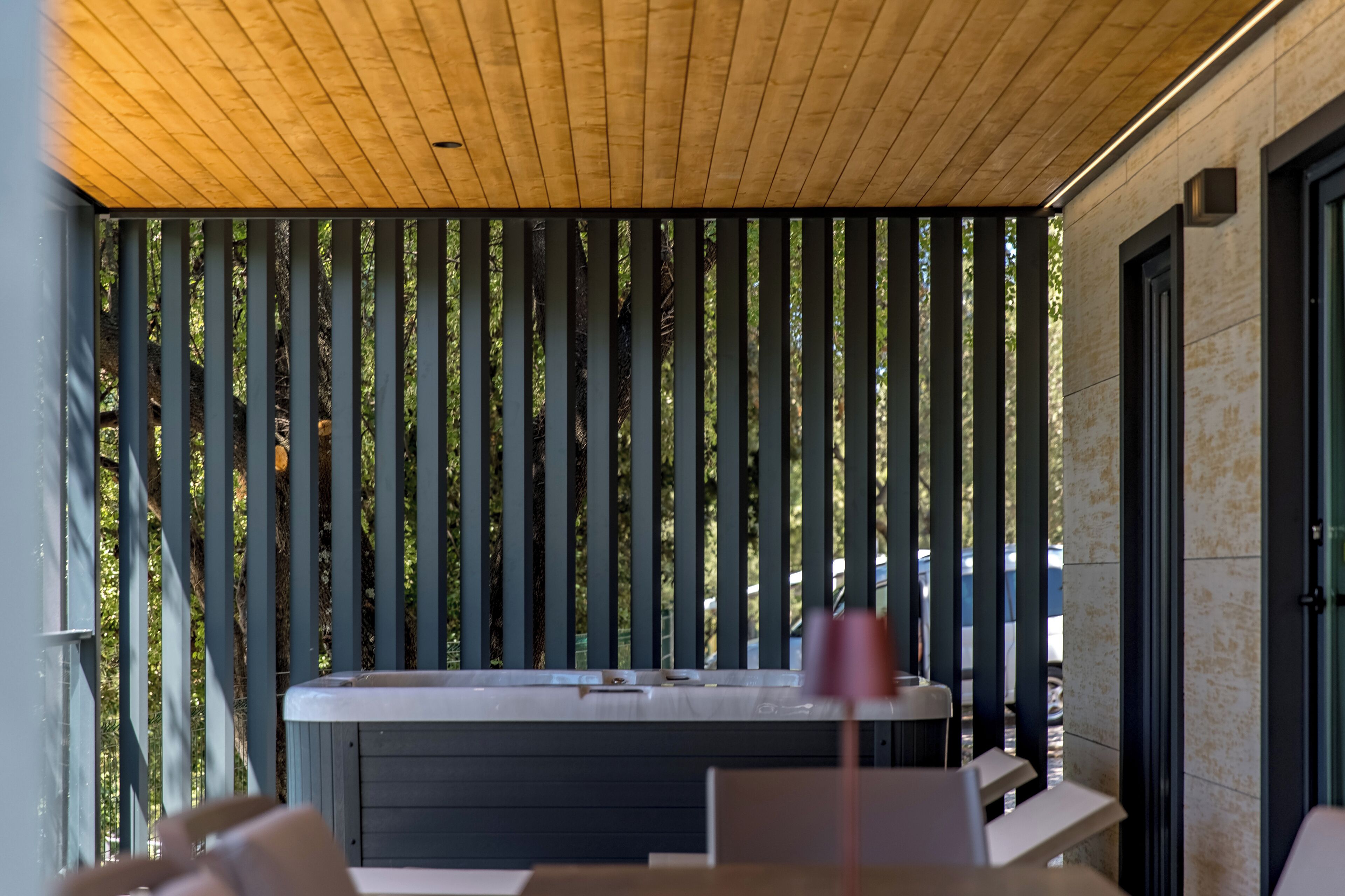 A modern outdoor seating area with a wooden ceiling and vertical blinds, a hot tub, and a stone wall.
