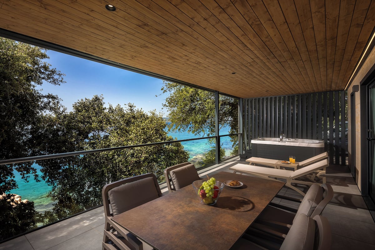 A modern outdoor dining area with a wooden ceiling, a large table with chairs, a fruit bowl, and a hot tub.