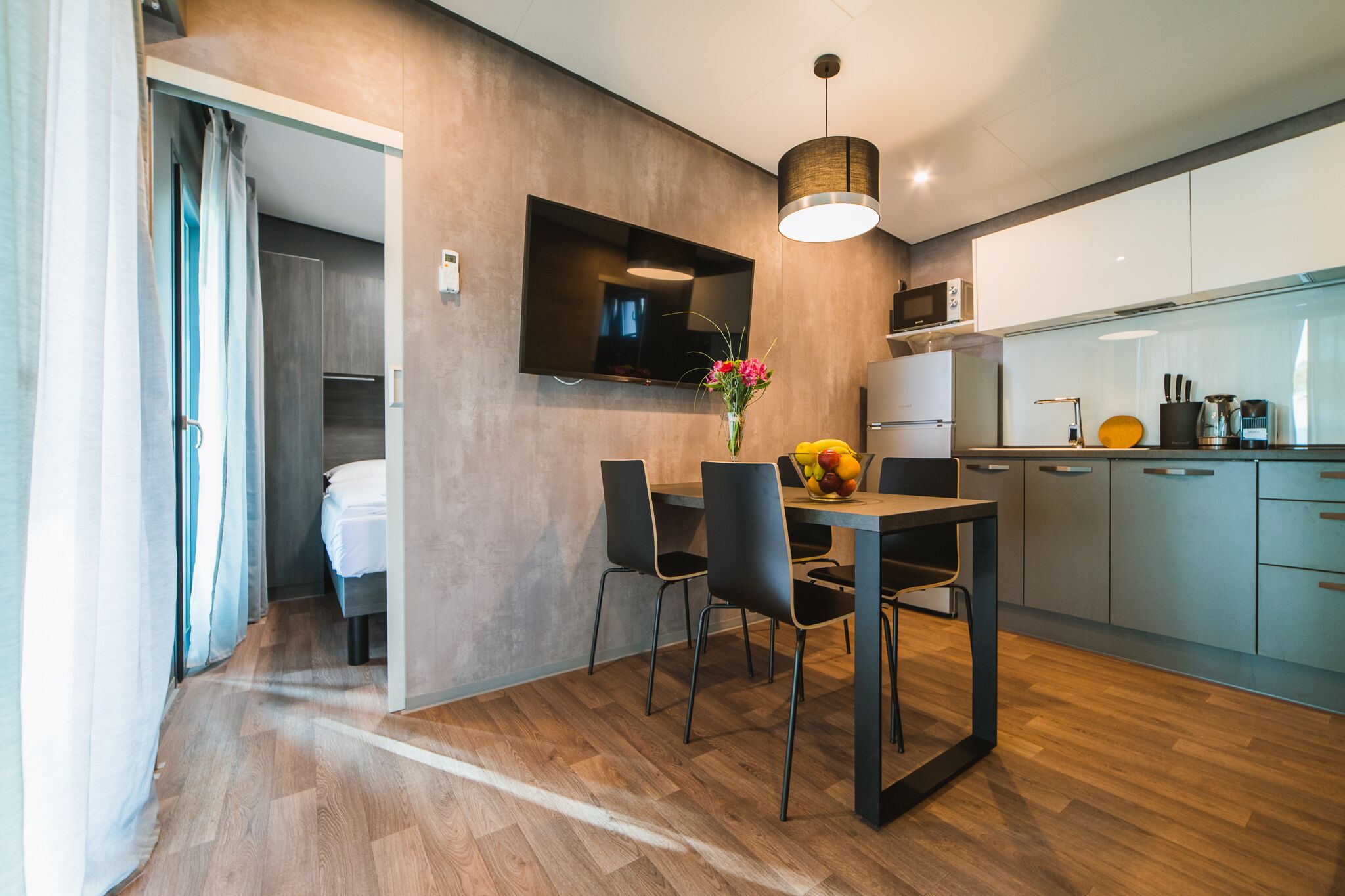 A modern kitchen with a dining area, wooden flooring, and a flat-screen TV mounted on the wall.