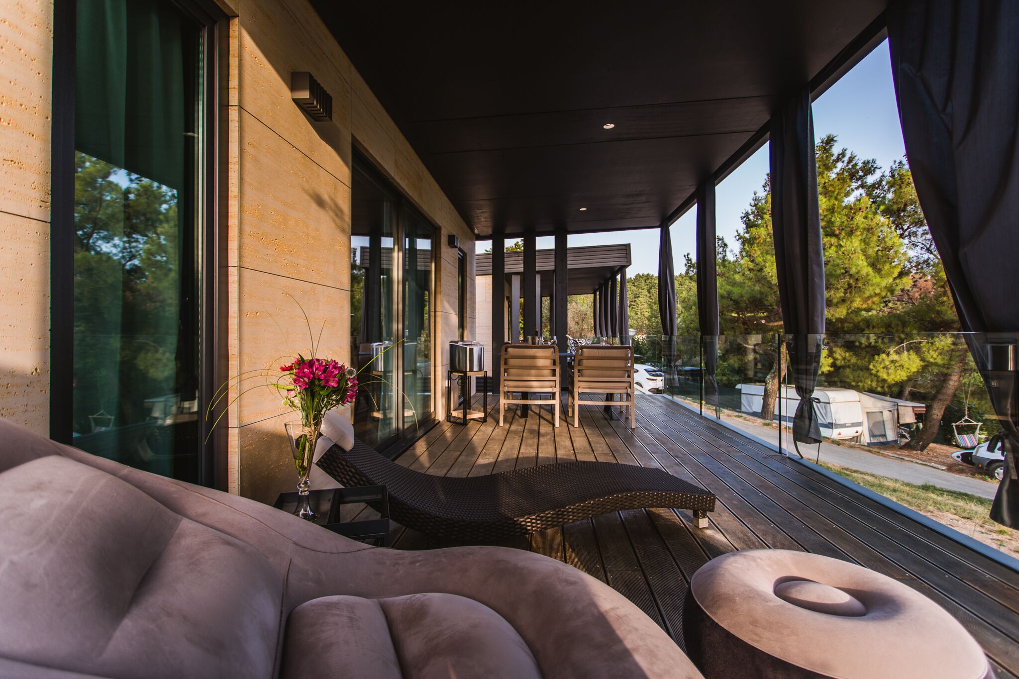 A modern outdoor seating area with a wooden deck, cushioned seating, and a glass wall providing a view of the surrounding landscape.