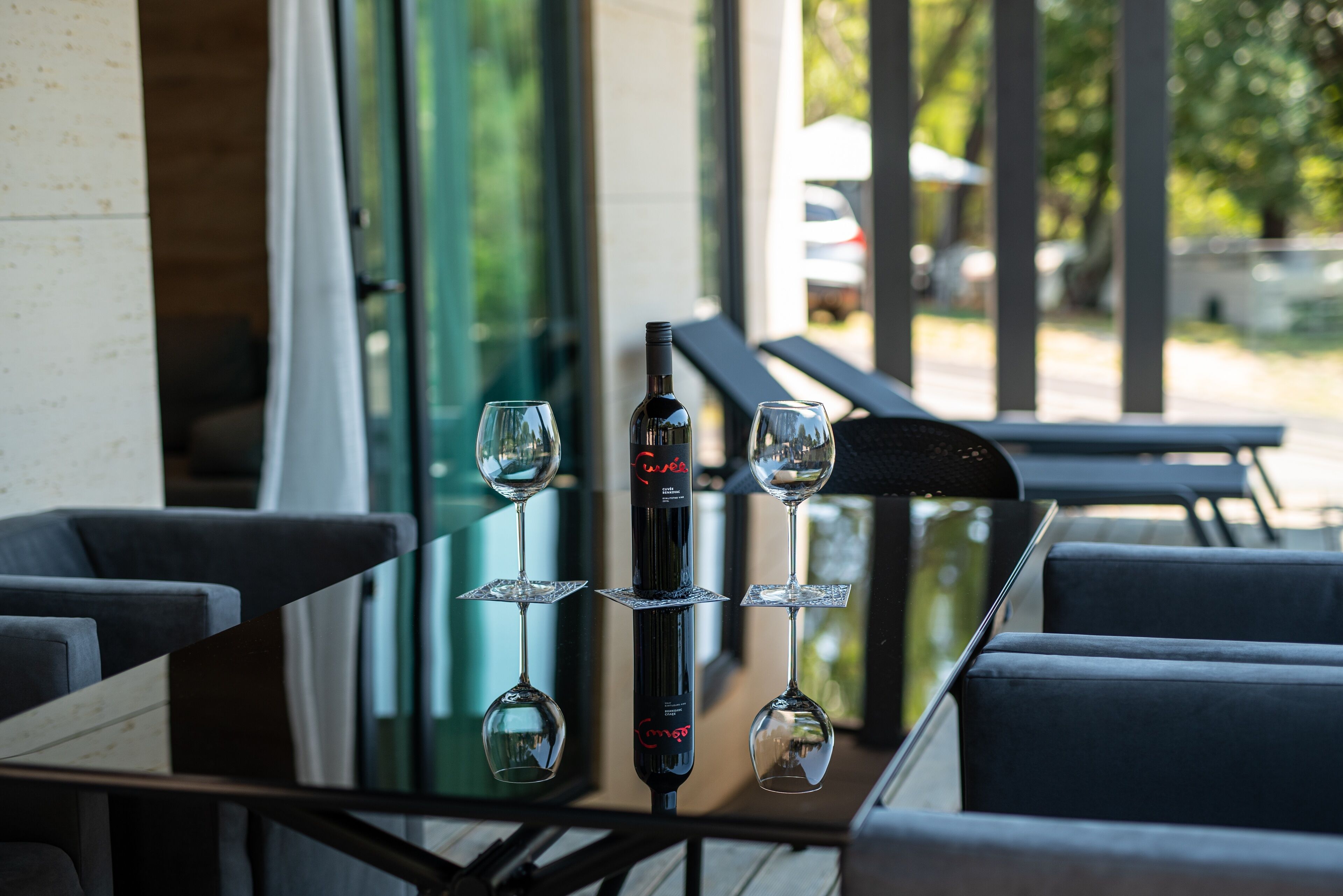 A table with a wine bottle and three wine glasses, set against a modern interior with large windows and a view of the outside.
