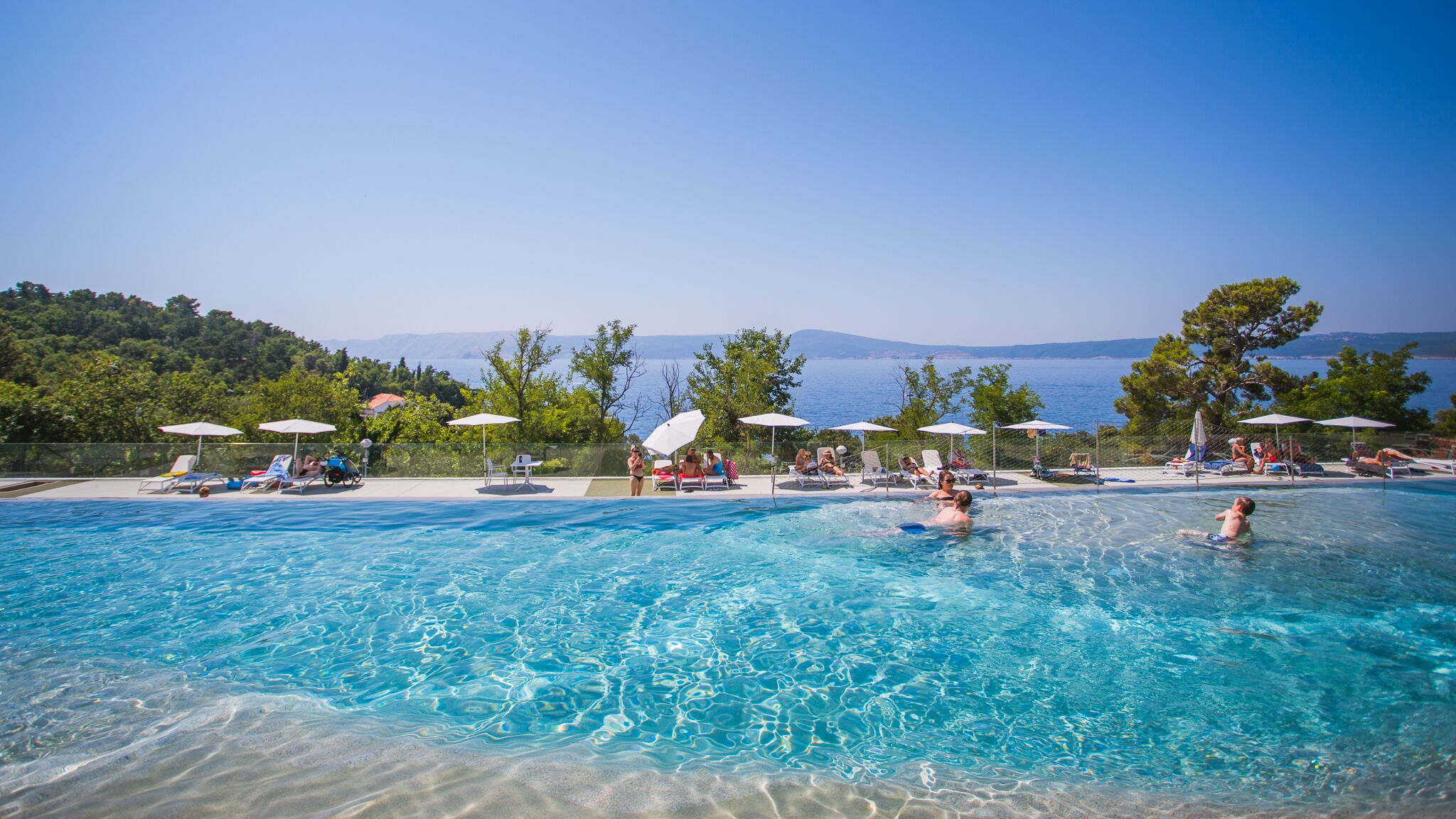 A pool area with clear blue water, lounge chairs, umbrellas, and a view of the sea and mountains.