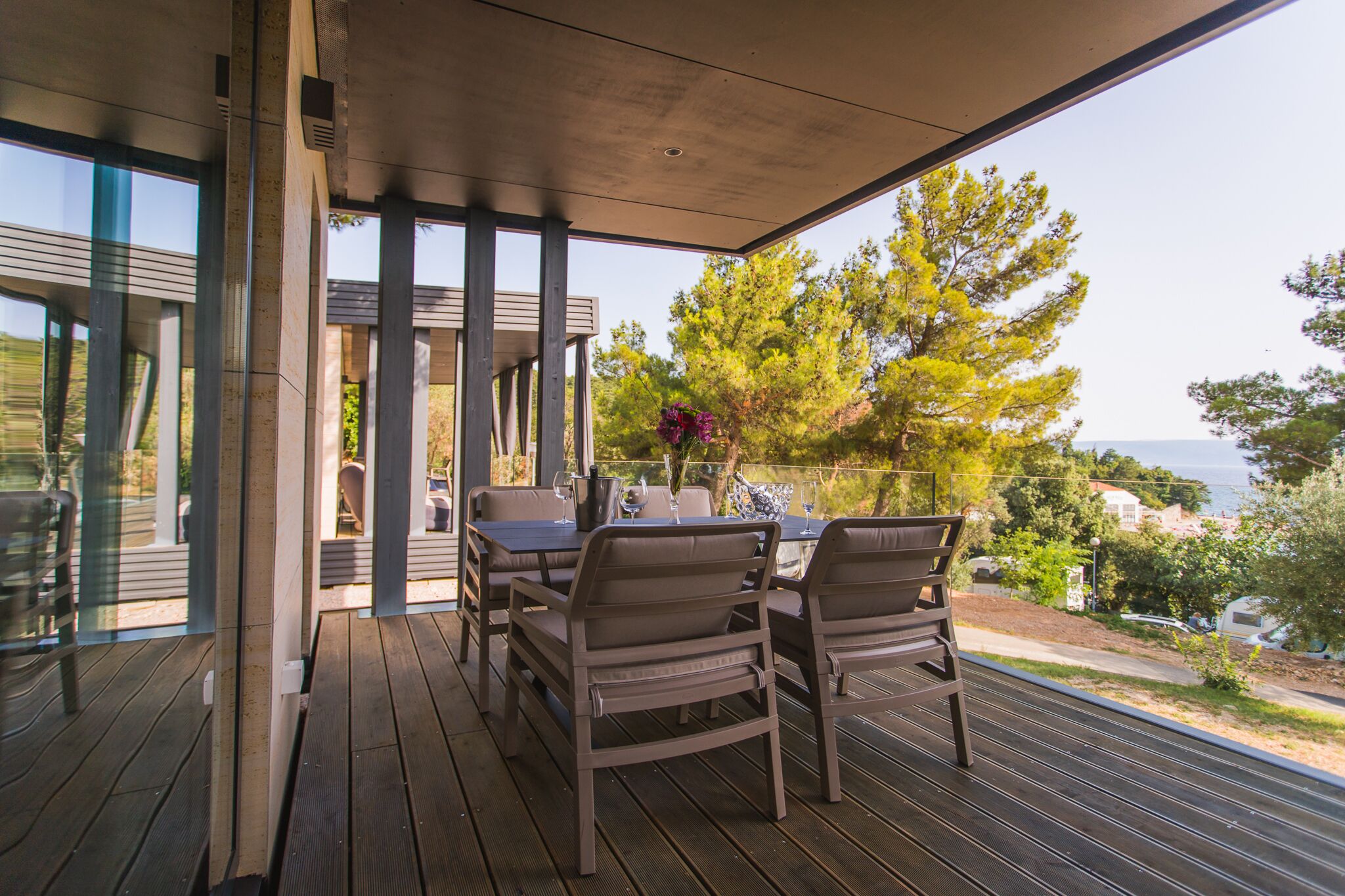 A covered outdoor dining area with a table and chairs, overlooking a scenic landscape with trees and a view of the sea.