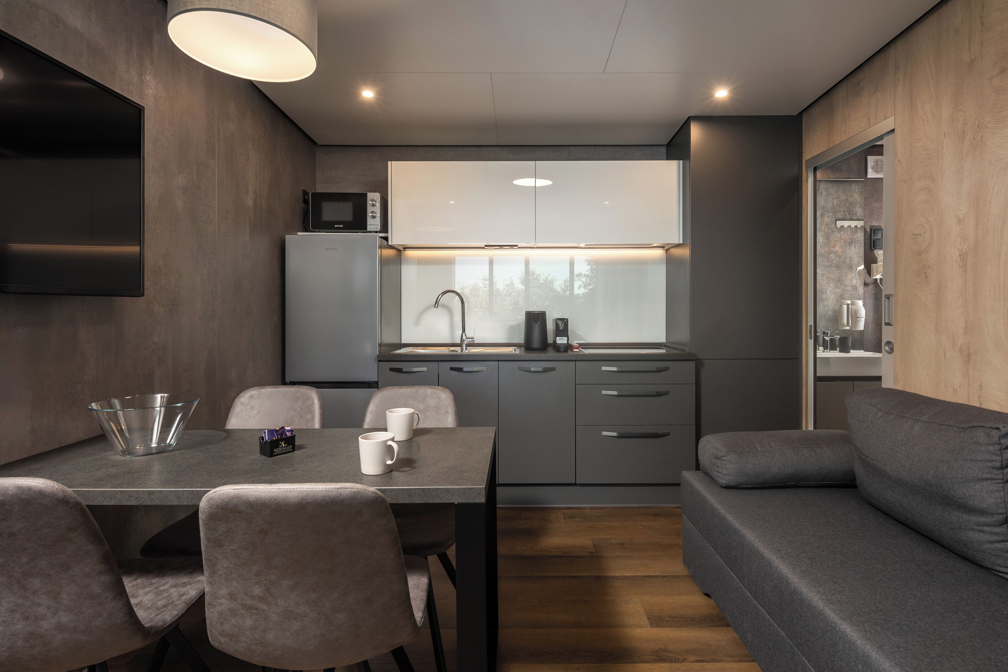 A modern kitchen with grey cabinets, a microwave, and a coffee maker. There is a dining table with chairs and a grey sofa.