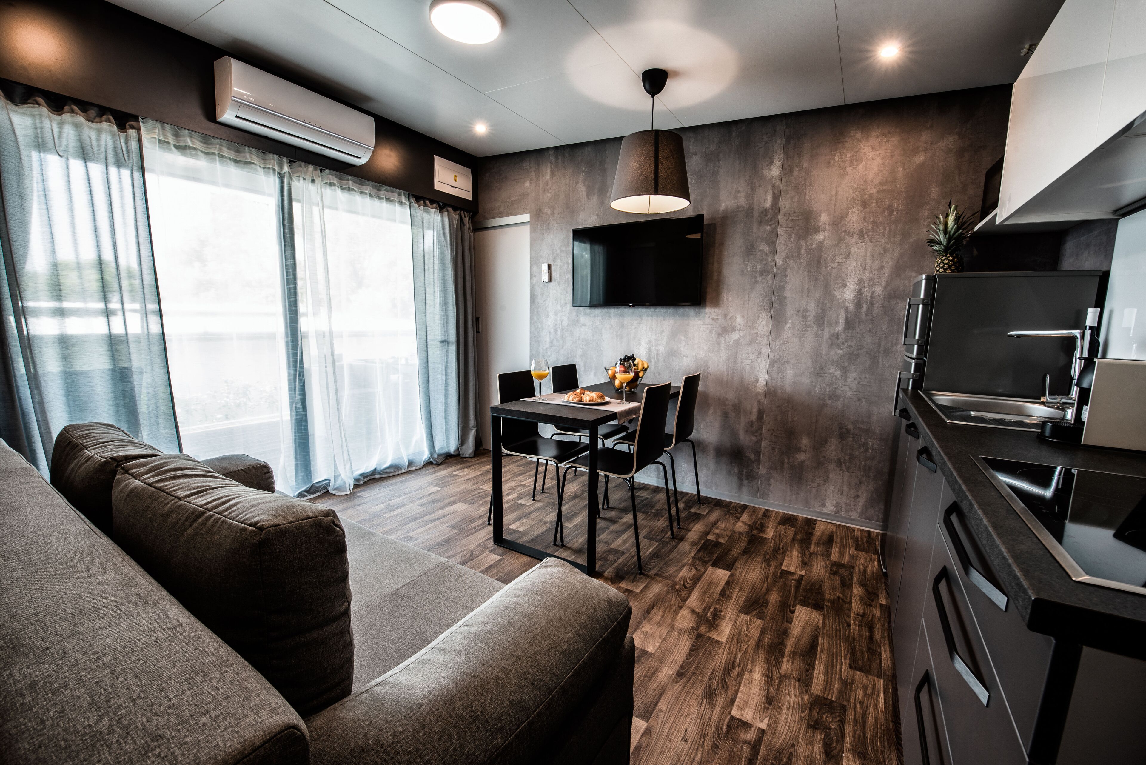 A modern kitchen with a dining area, wooden flooring, and a large window with curtains.
