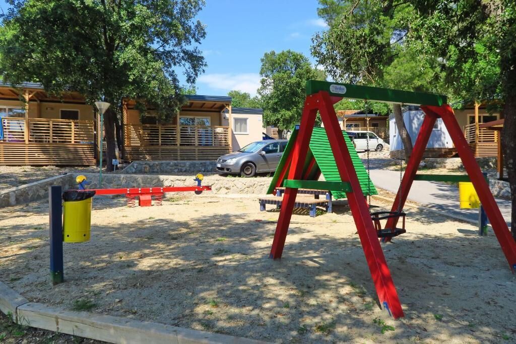 Un parc infantil amb balancins i un tròlit, envoltat d'arbres i un edifici al fons.
