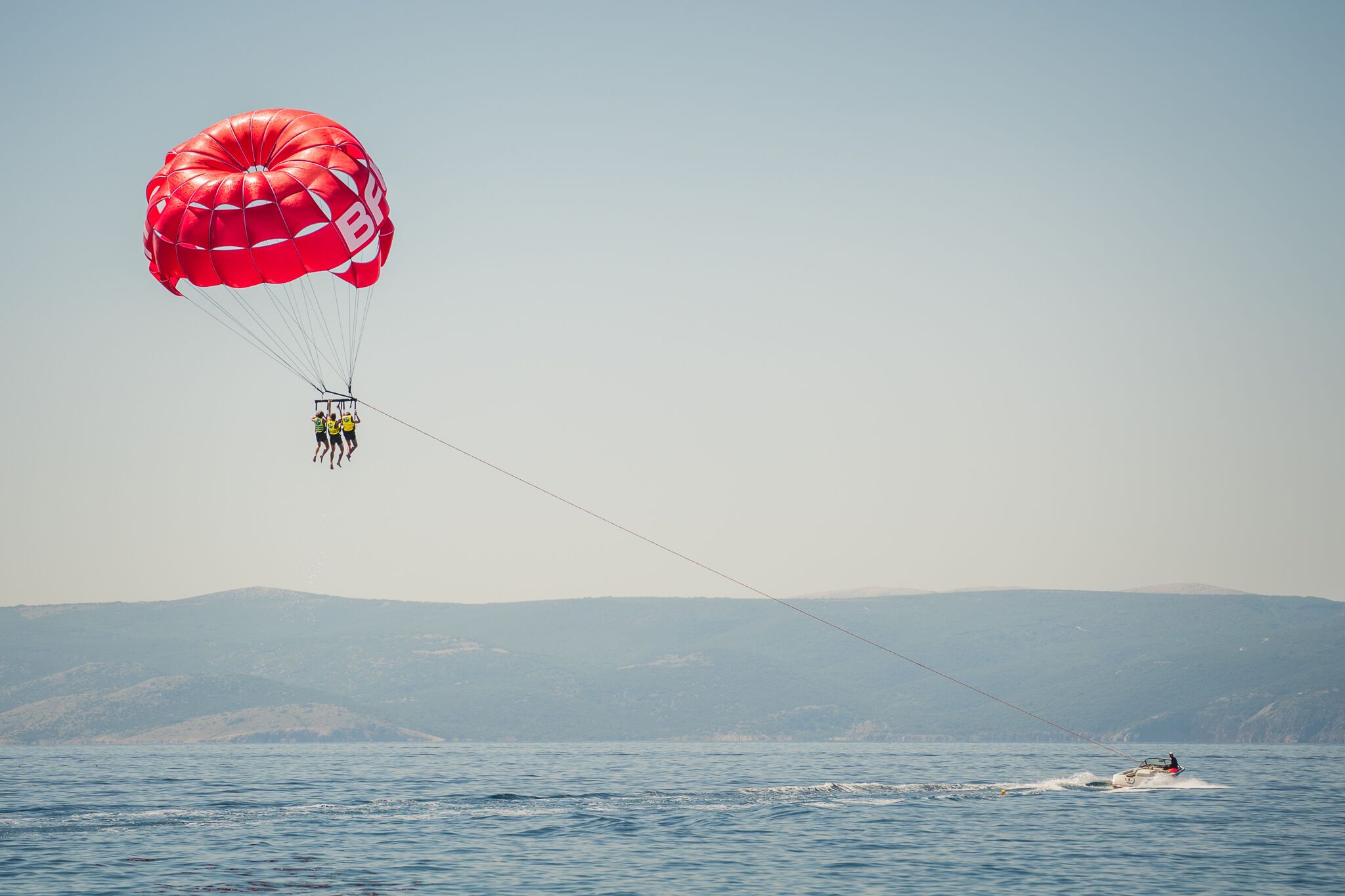 A parasailer with a red canopy and a person in a yellow life jacket, being towed by a speedboat on a body of water with distant hills.