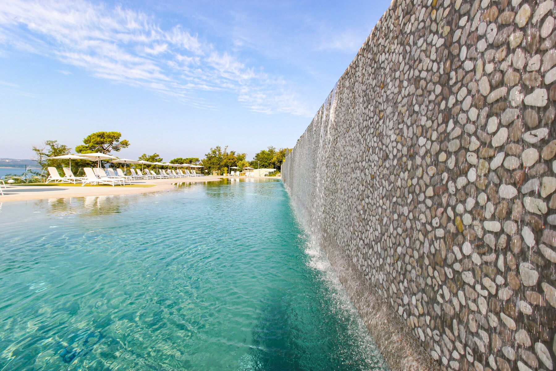 A clear pool with a pebble wall, lounge chairs, and a view of the beach.