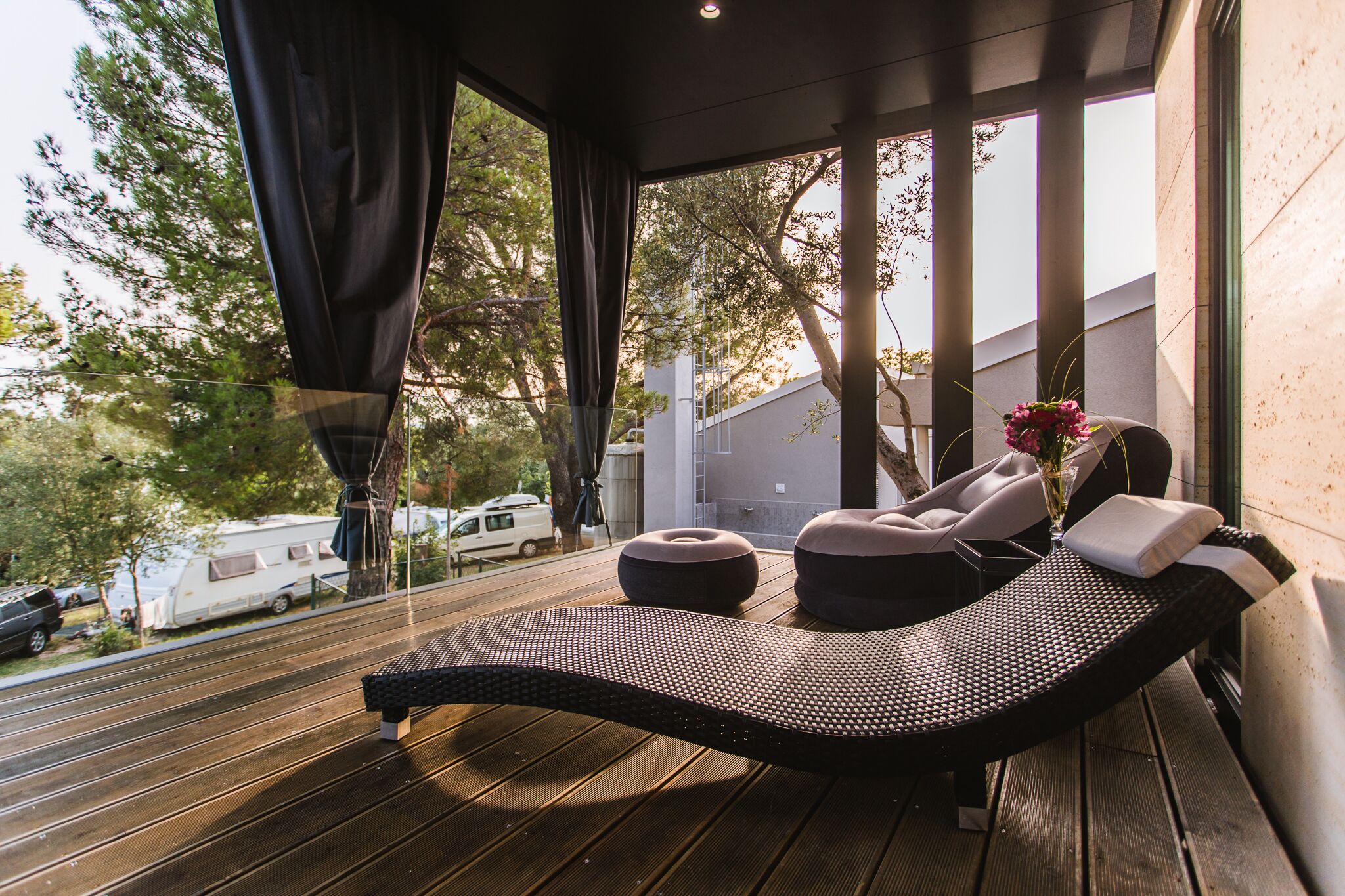 A sun lounger with cushions and a vase of flowers, a wooden deck, and a view of trees and a white van in the background.