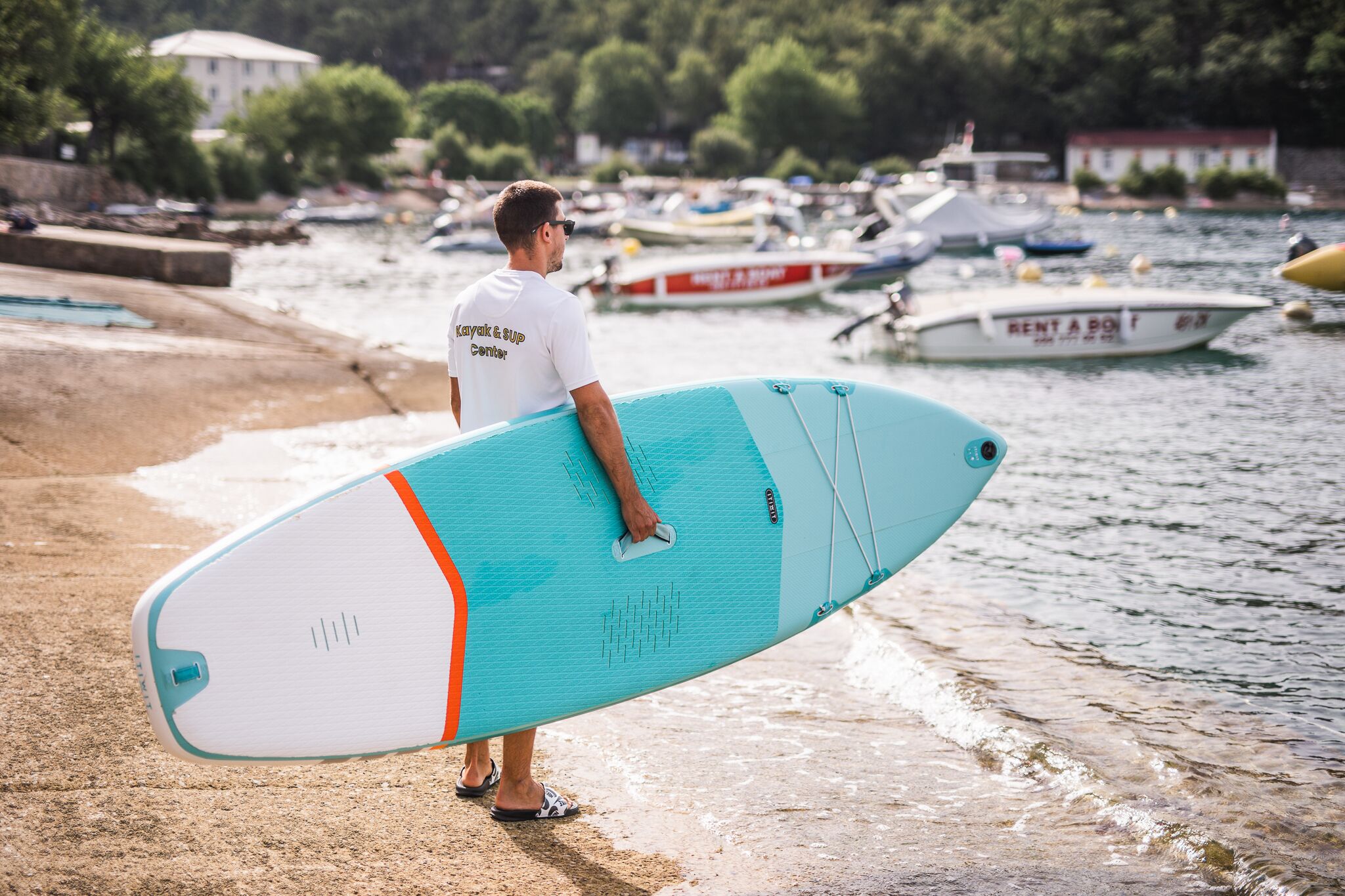 Une personne tenant une planche de surf sur une plage, avec des bateaux en arrière-plan.