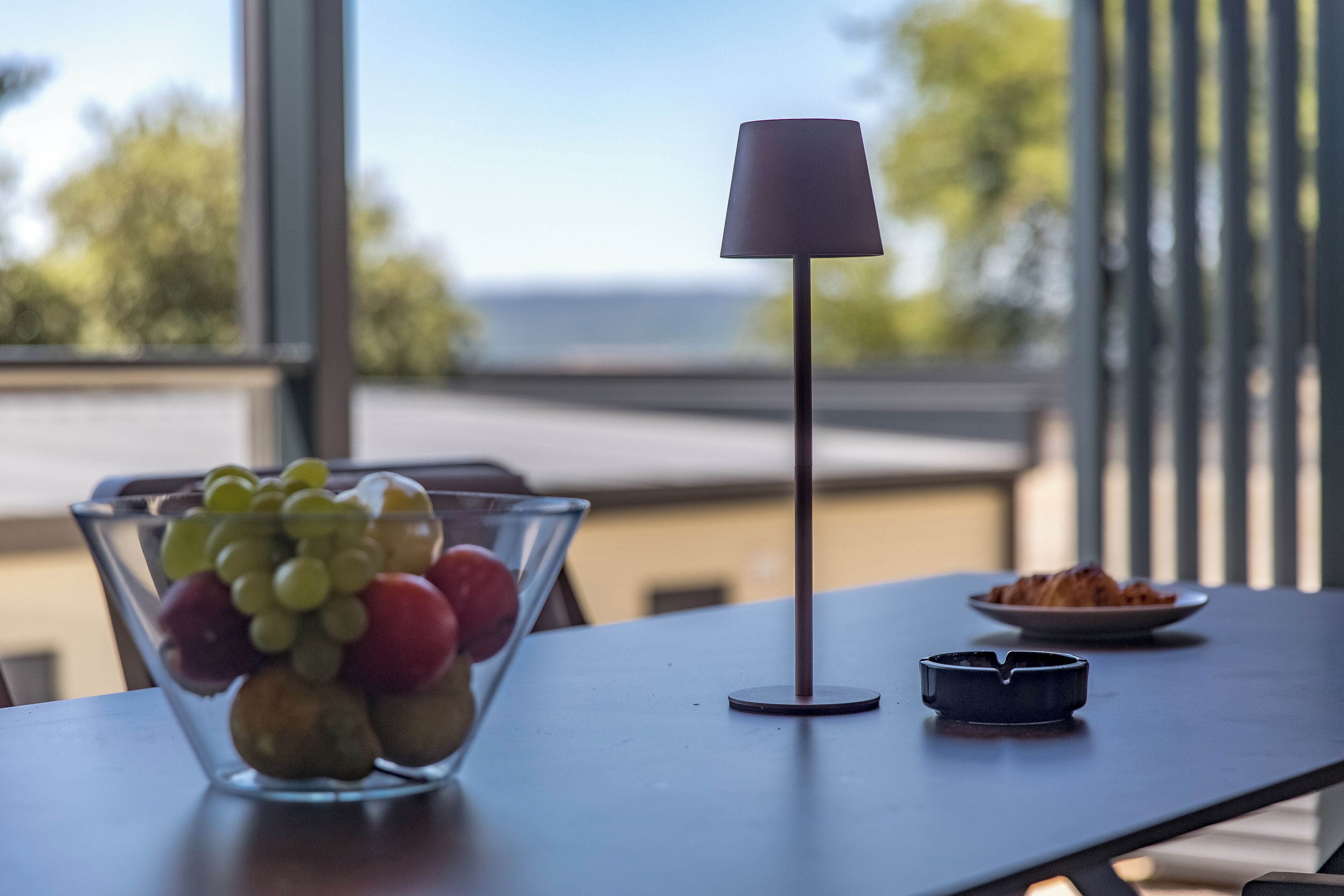 A table with a bowl of fruit, a lamp, and a plate of food.